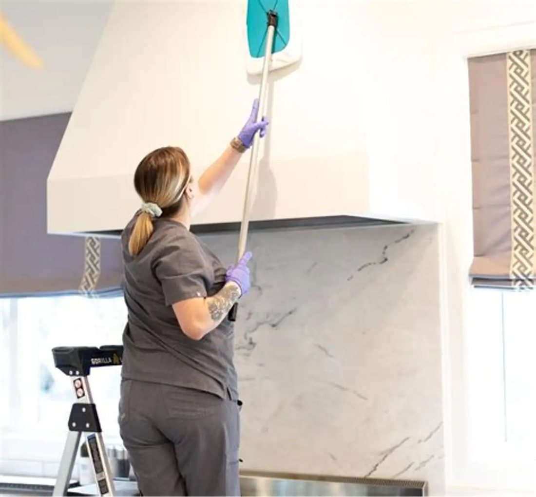 Cleaner in gloves wiping down a kitchen range hood with an extension pole